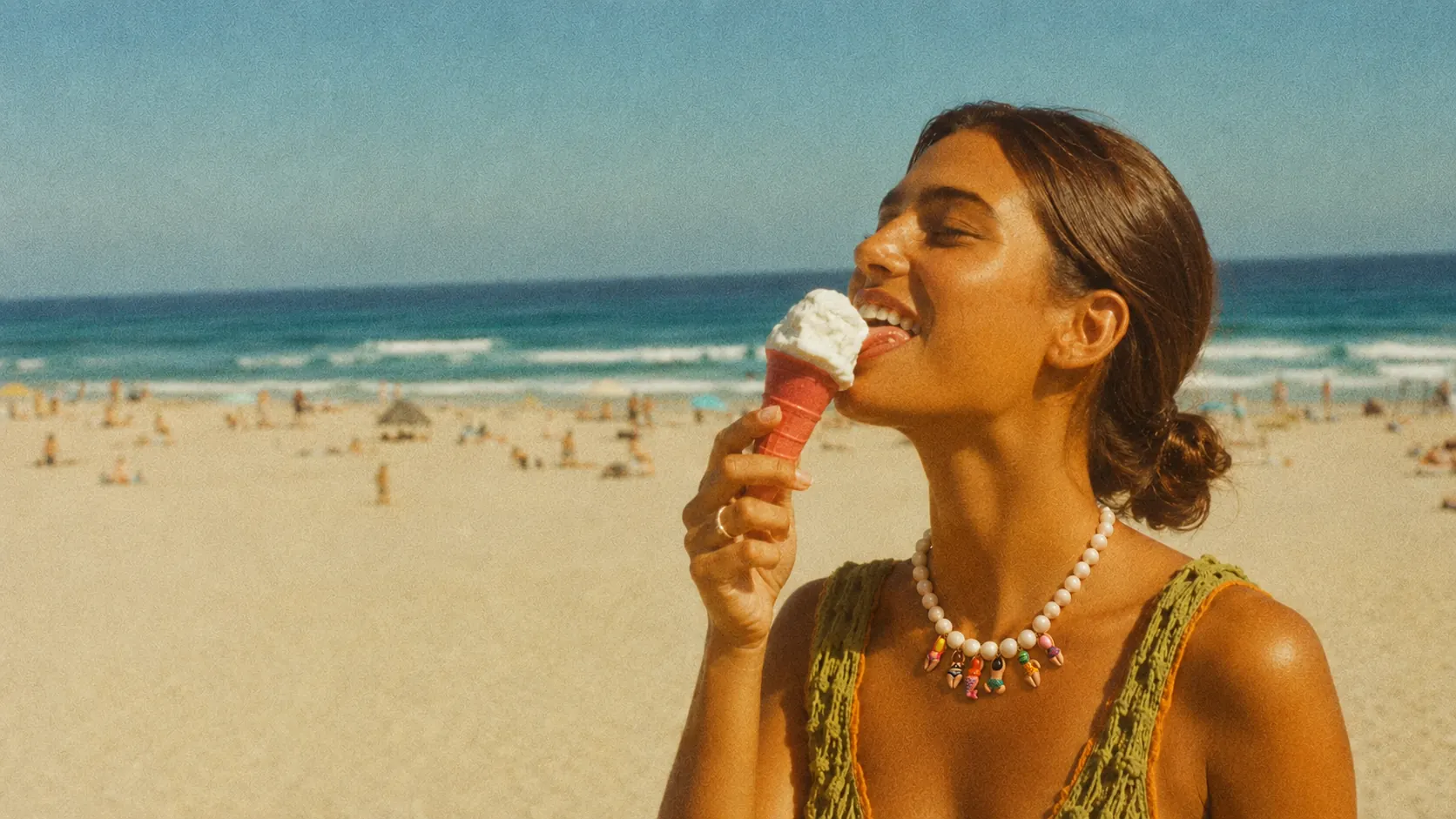 Femme souriante sur une plage ensoleillée, portant un collier de perles blanches à figurines colorées, une glace à la main